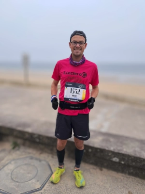 Nicolas, vêtu d'un t-shirt de course rose et portant un dossard, se tient prêt à participer au Marathon de la Côte d'Amour 2024, avec la plage en arrière-plan.