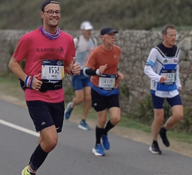 Un coureur portant un t-shirt rose et des lunettes, souriant pendant sa course, entouré d'autres participants sur une route bordée de verdure et d'un mur de pierre.