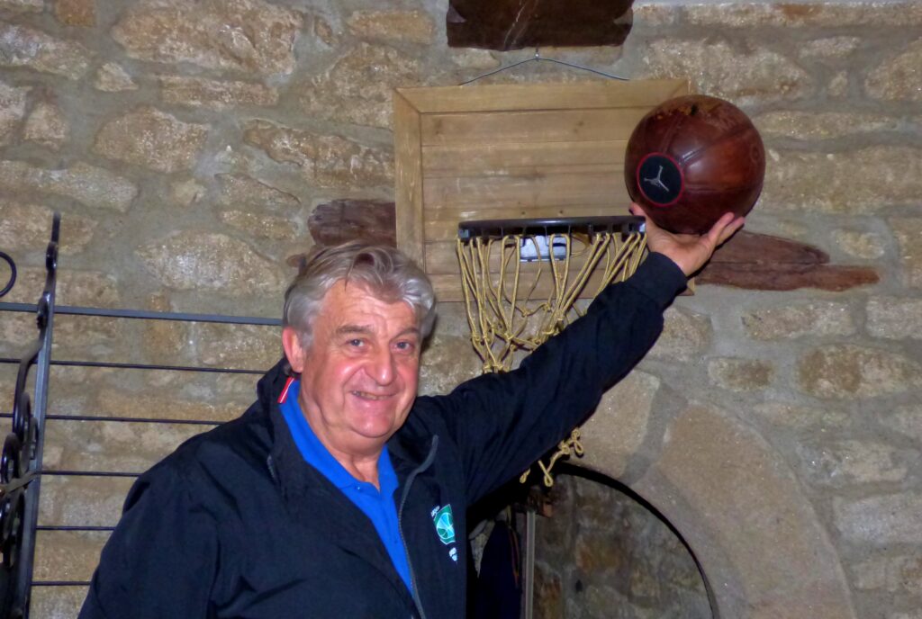 : Homme souriant tenant un ballon de basketball en hauteur devant un panier et un mur en pierre.