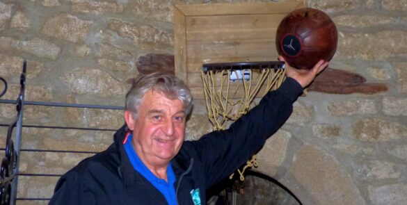 : Homme souriant tenant un ballon de basketball en hauteur devant un panier et un mur en pierre.