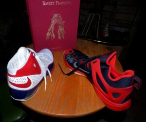 Deux chaussures de basketball rouges et noires posées sur une table en bois, devant un livre intitulé "Basket Français