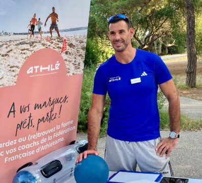 Un éducateur sportif souriant, vêtu d'un t-shirt bleu, présente du matériel d'entraînement sport-santé lors d'un stand en extérieur.