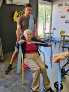 Un éducateur sportif guide un groupe de seniors dans un exercice de renforcement et d'équilibre avec des bâtons, dans une salle adaptée.