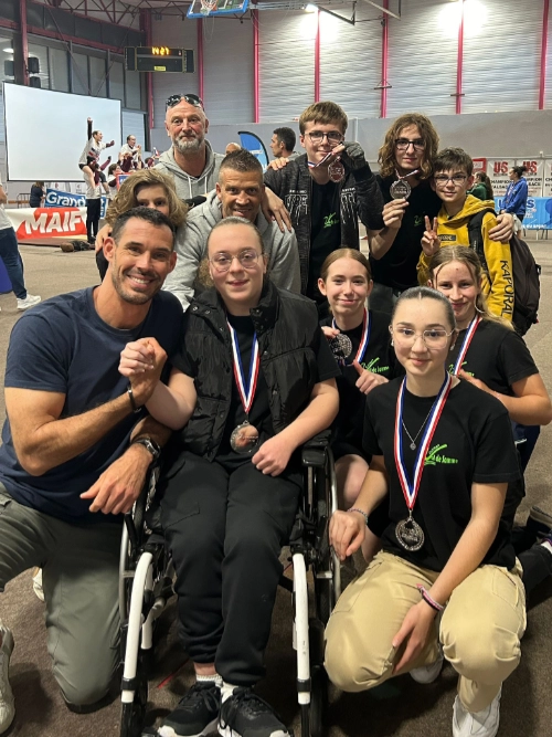 Un groupe de jeunes athlètes, dont une en fauteuil roulant, pose fièrement avec leurs médailles aux côtés de leur éducateur sportif dans une salle de compétition.