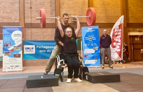 Une jeune athlète en fauteuil roulant soulève une barre d'haltérophilie avec l'assistance de son éducateur, dans le cadre d'une compétition inclusive.