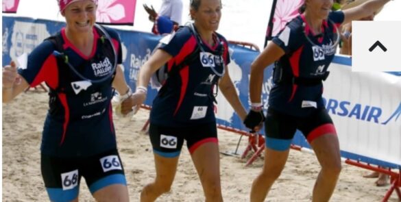 Trois femmes souriantes courent main dans la main sur une plage lors d'un trail solidaire, portant des dossards numérotés et des tenues de sport assorties. Elles incarnent l'esprit d'équipe et la détermination. En arrière-plan, on distingue des spectateurs et des bannières promotionnelles. Cet événement est lié au "Trail Tous avec Alex 2025" organisé à Pornichet.