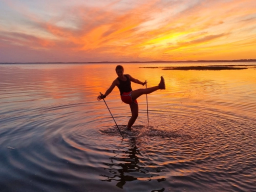 Pratiquante de marche nordique effectuant un mouvement technique dans l’eau au coucher du soleil, illustrant une approche créative de la marche nordique technique et pédagogie.