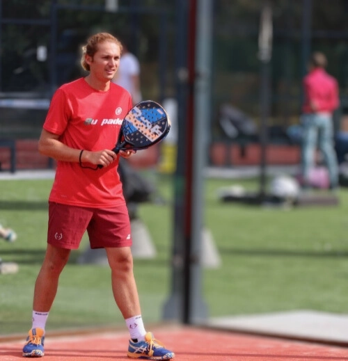 Joueur de padel en tenue rouge sur un terrain extérieur, raquette en main, prêt à engager un échange.