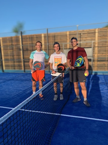 Trois joueurs de padel en plein air posent sur un terrain bleu, raquettes à la main, prêts pour un match.