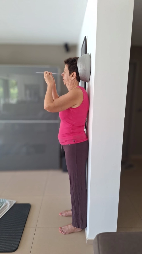 Femme debout contre un mur avec un coussin derrière la tête, pratiquant un exercice postural d’éducation corporelle pour soulager la douleur.