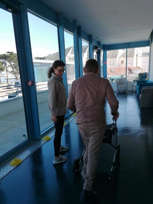 Inès accompagne Michel qui marche avec un déambulateur pendant une séance d’activité physique adaptée dans un centre de rééducation