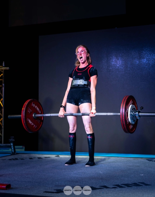 Athlète féminine en force athlétique réalisant un soulevé de terre sur une plateforme, avec barre chargée et expression d’effort intense.