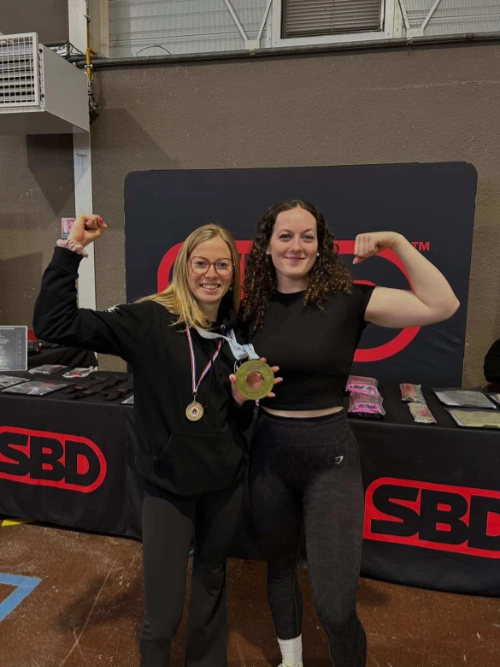 Deux athlètes féminines de force athlétique posent en salle de compétition, médailles au cou, en flexion de biceps devant un stand SBD.