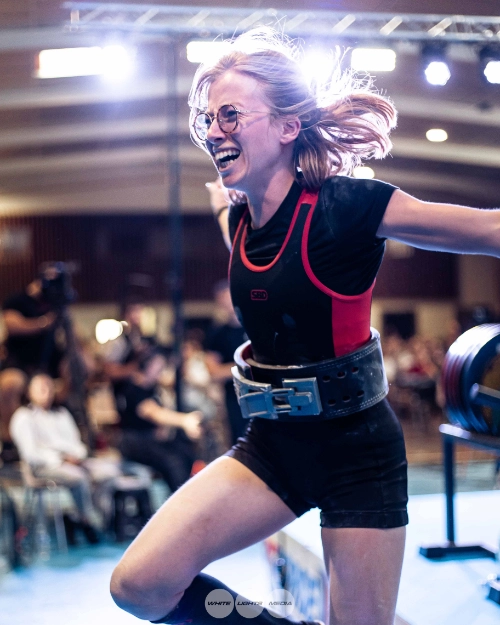 Athlète féminine en force athlétique célébrant sa performance sur une plateforme de compétition, bras ouverts et sourire, avec ceinture de musculation.