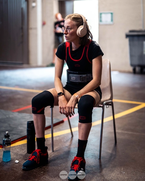 Athlète féminine en force athlétique assise sur une chaise en salle de compétition, portant une ceinture de musculation, des genouillères et un casque audio.