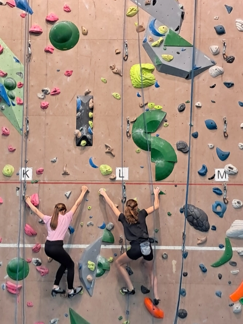 Deux jeunes filles grimpent côte à côte sur un mur d’escalade en salle avec des prises colorées, dans un club d’escalade à Saint-Molf.