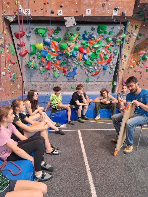 Groupe de jeunes assis dans une salle d’escalade, écoutant un encadrant devant un mur de bloc, dans un club d’escalade à Saint-Molf.