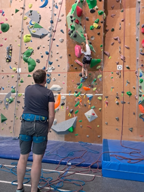 Jeune grimpeuse sur un mur d’escalade en salle, observée depuis le sol par une autre personne, dans un club d’escalade à Saint-Molf.