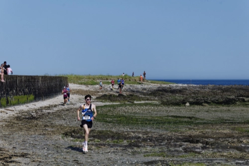 Coureur de la Course Nature du Croisic 2026 sur un sentier côtier rocheux, avec d’autres participants et l’océan en arrière-plan.
